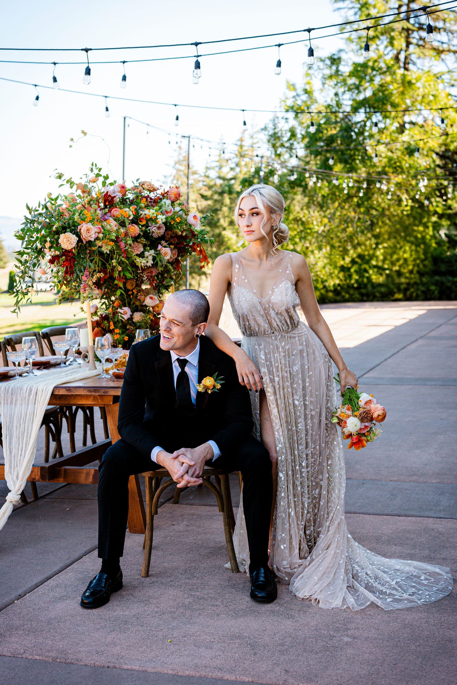 Elegant wedding couple poses at outdoor reception with string lights and floral arrangements on a patio setting.