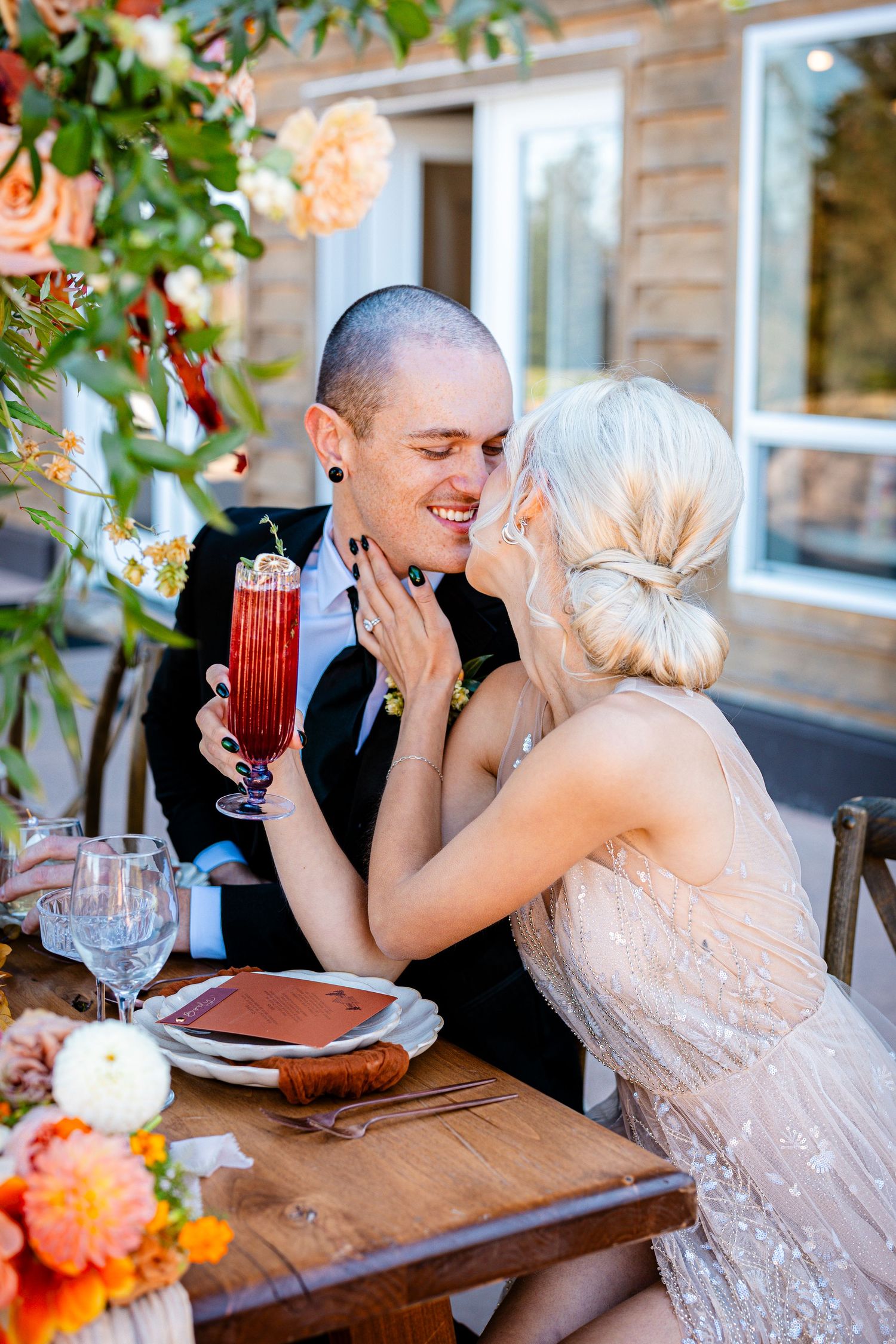Couple shares intimate moment at rustic outdoor wedding reception with colorful floral decor and red cocktail.