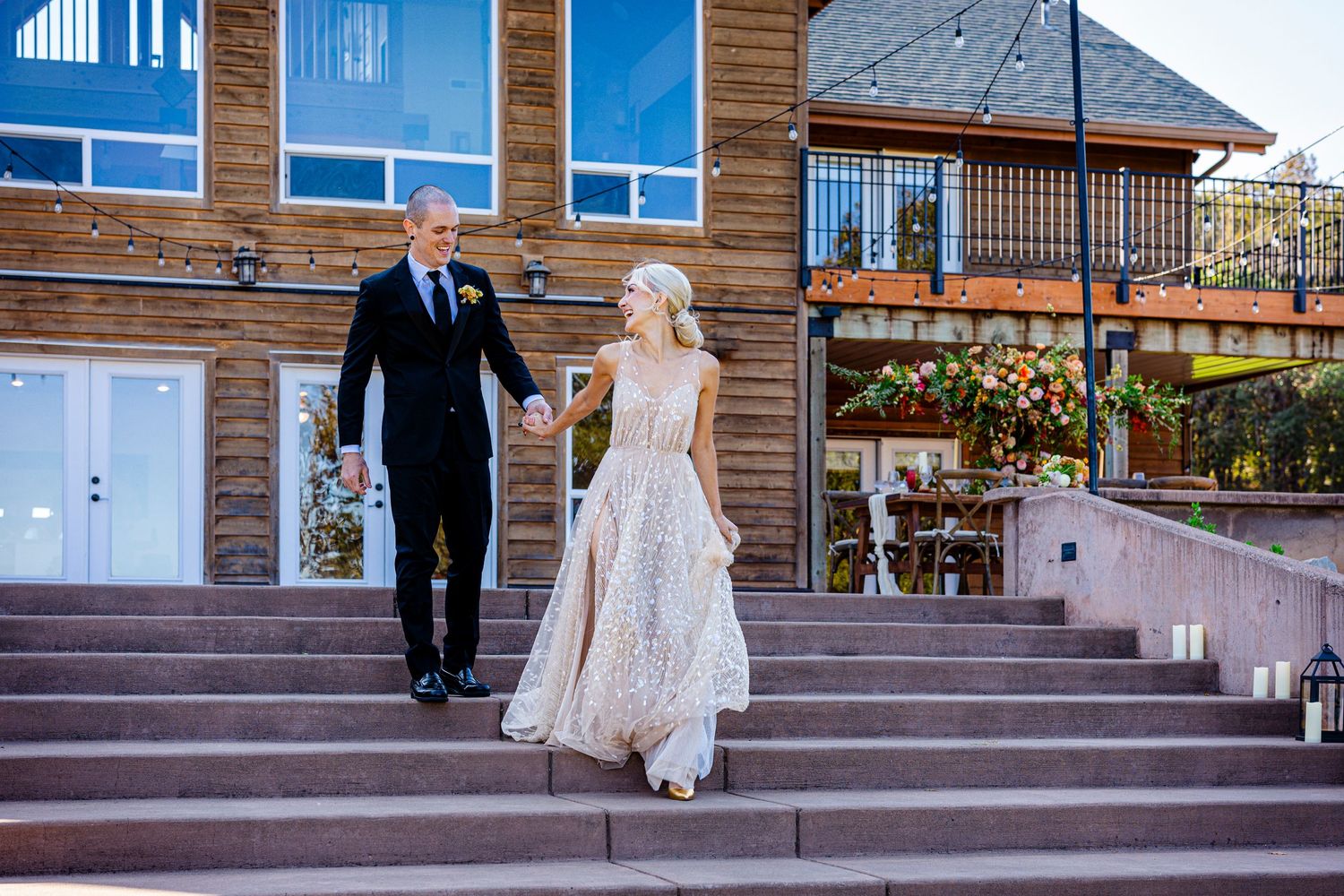 A couple in formal wedding attire descends stone steps outside a rustic wooden building with blue windows.