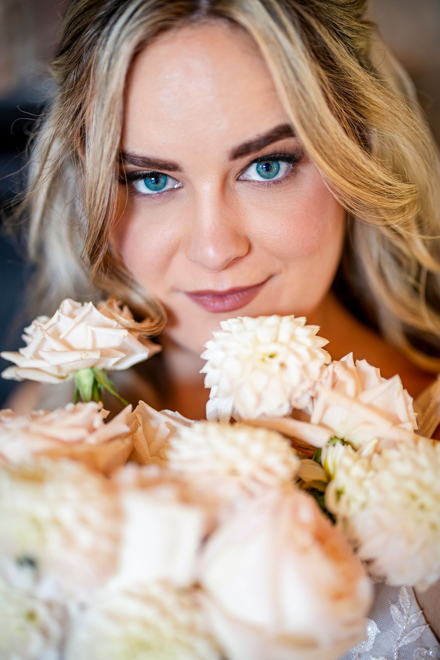 A close-up portrait with soft peach-colored roses in the foreground, highlighting striking blue eyes and blonde hair.