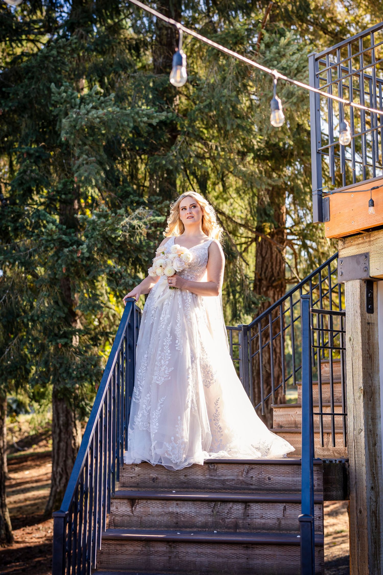 A person in a white lace wedding gown stands on outdoor stairs with string lights and trees in the background.