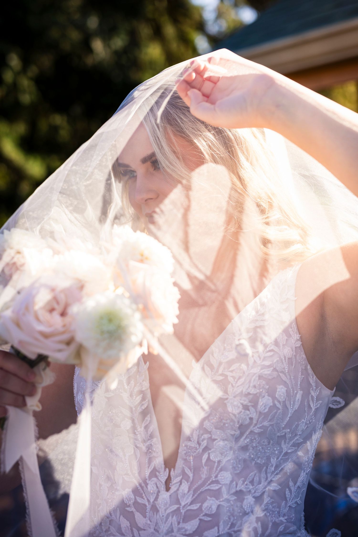 A bride wearing a delicate lace dress and veil holds a soft pink and white bouquet in sunlit outdoor setting.