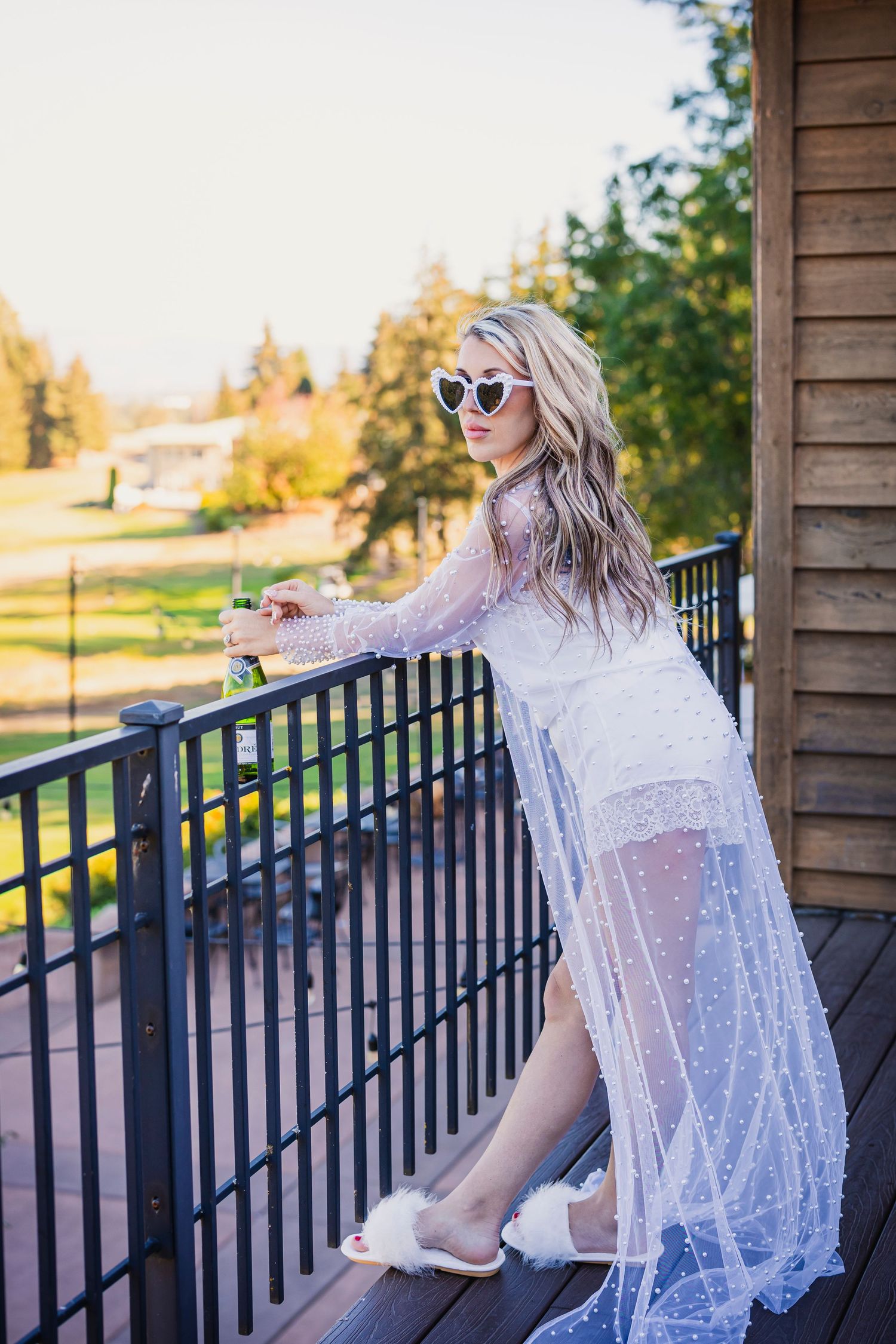 Woman in white lace dress and sunglasses poses on wooden deck railing during golden sunset.