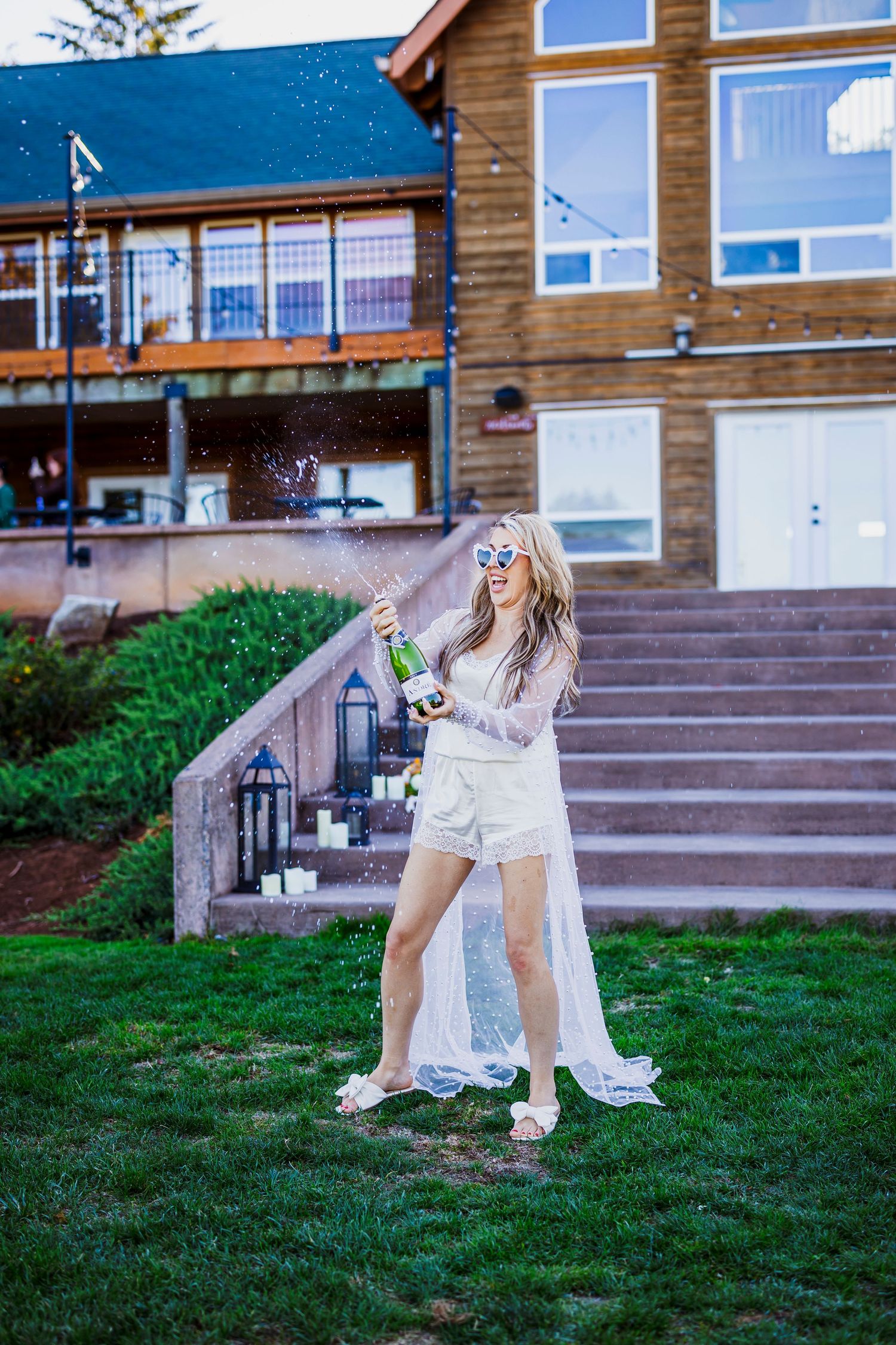 A bride in a flowing white dress and cape stands on a lawn in front of a rustic wooden lodge.