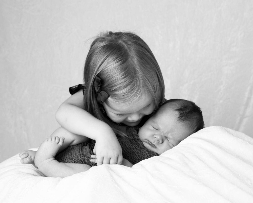 Nyfødt baby liggende på et laken i fotostudio med storesøster som koser bakfra på nyfdøtfotografering i Lyngdal