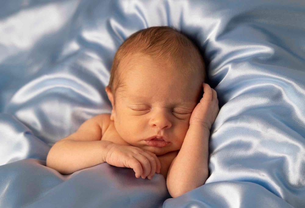 Portrett av nyfødt baby gutt liggende i silke og sover med hånden på kinnet på nyfødtfotografering i Lyngdal