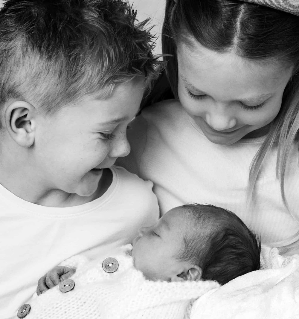 To søsken som holder nyfødtbaby som sover i armene, på nyfødtfotografering i Lyngdal