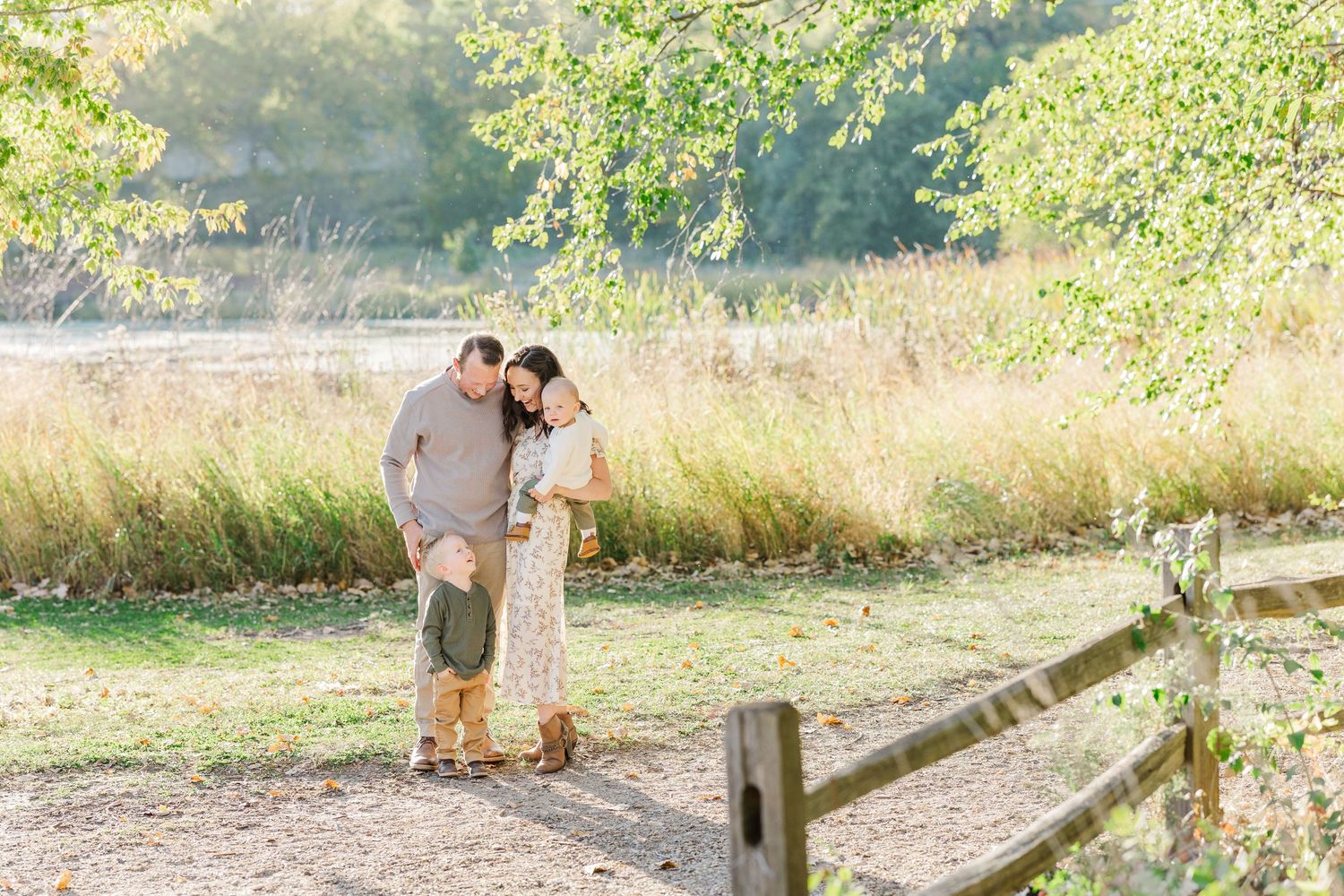 A couple stands together on a sunlit nature trail surrounded by tall grass and trees on an autumn day.