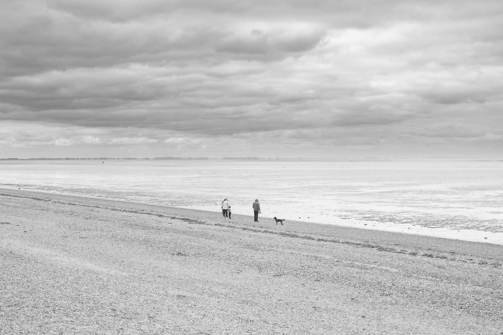 Three silhouetted figures walk along a sloping beach against a dramatic cloudy sky in black and white.