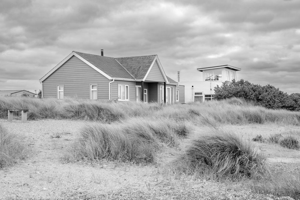 A black and white beach house sits among tall grass dunes under a cloudy sky.