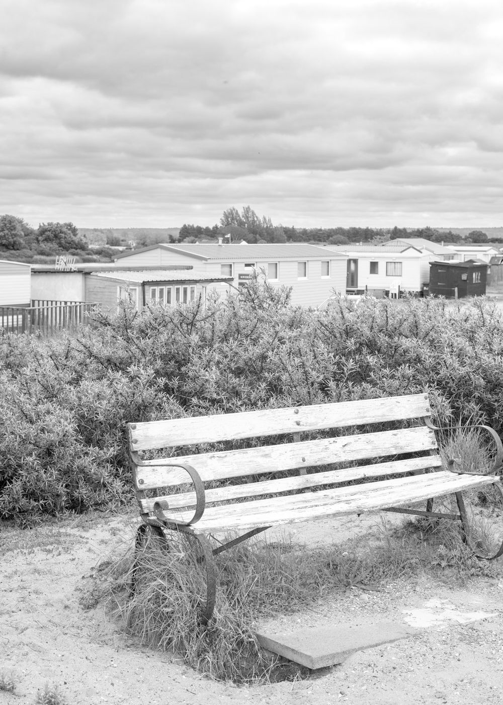 A weathered wooden bench sits in front of mobile homes on an overcast day, captured in black and white photography.