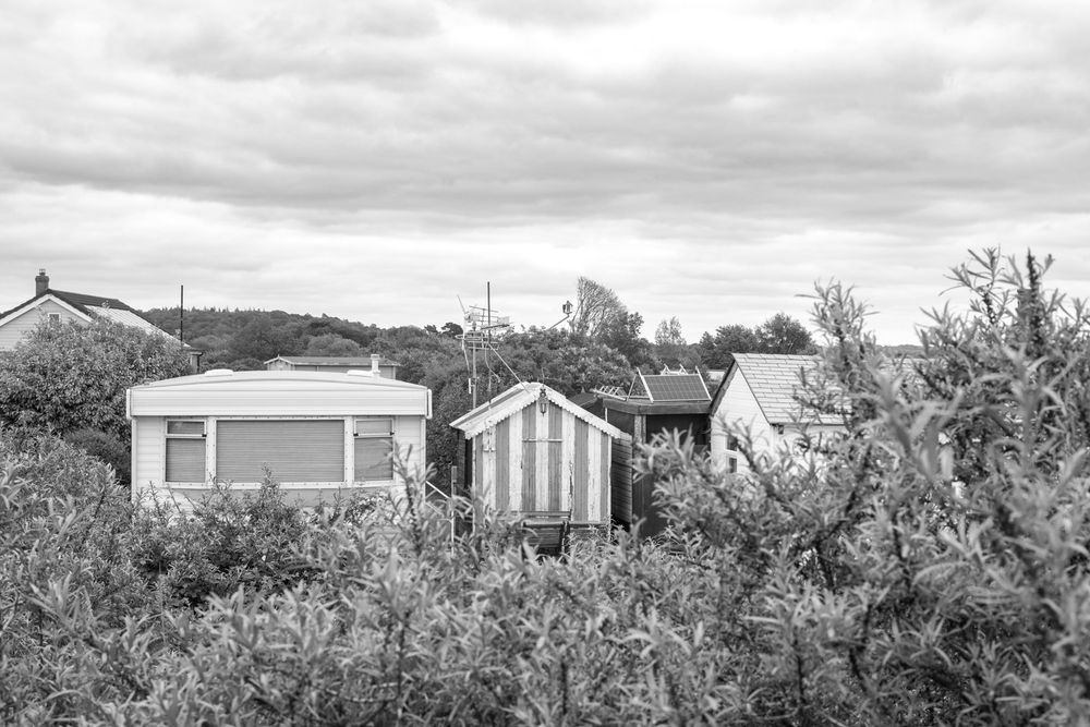 Black and white photo of small beach huts and seaside cottages nestled among coastal vegetation under cloudy skies.
