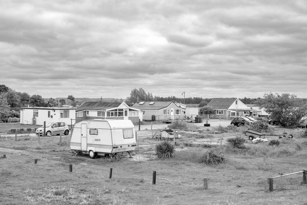 Black and white photo of a vintage caravan park with mobile homes and houses on a grassy landscape under cloudy skies.
