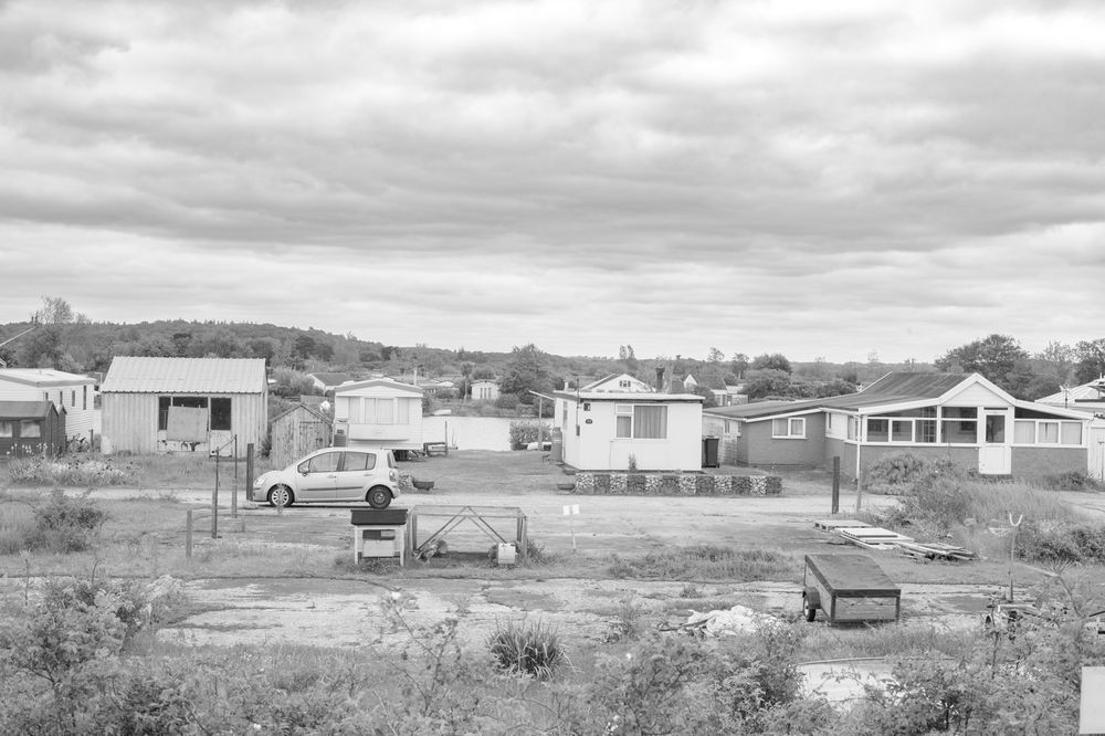 Black and white photograph of rural houses and cars scattered across undeveloped land with cloudy skies overhead.