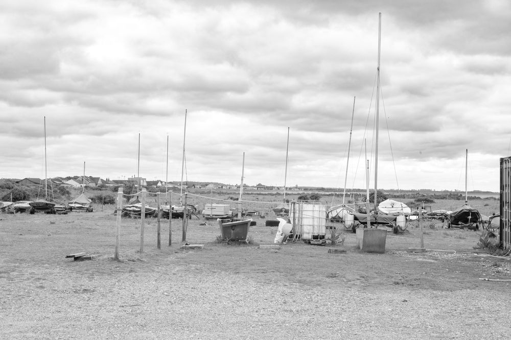 A black and white photograph shows sailboat masts and fishing equipment scattered across a dusty harbor yard.