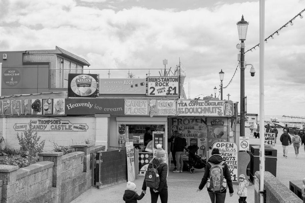 Vintage black and white photo of seaside boardwalk with shops and lampposts along a beachfront promenade.
