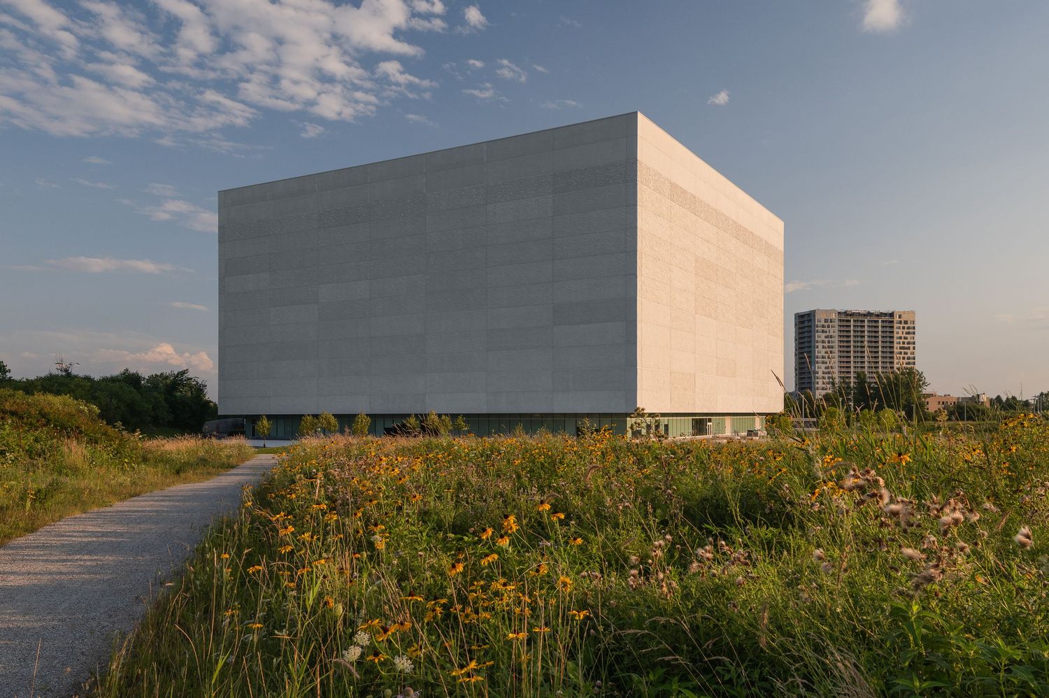 Minimalist white concrete building stands in a wildflower meadow against blue sky with wispy clouds.