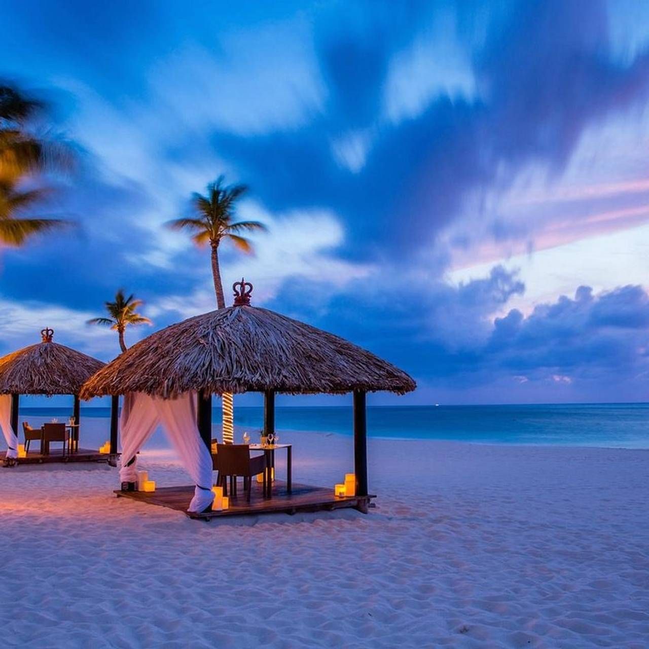 Palm trees and thatched beach cabanas illuminate the shoreline at sunset with dramatic purple and blue clouds over turquoise waters.