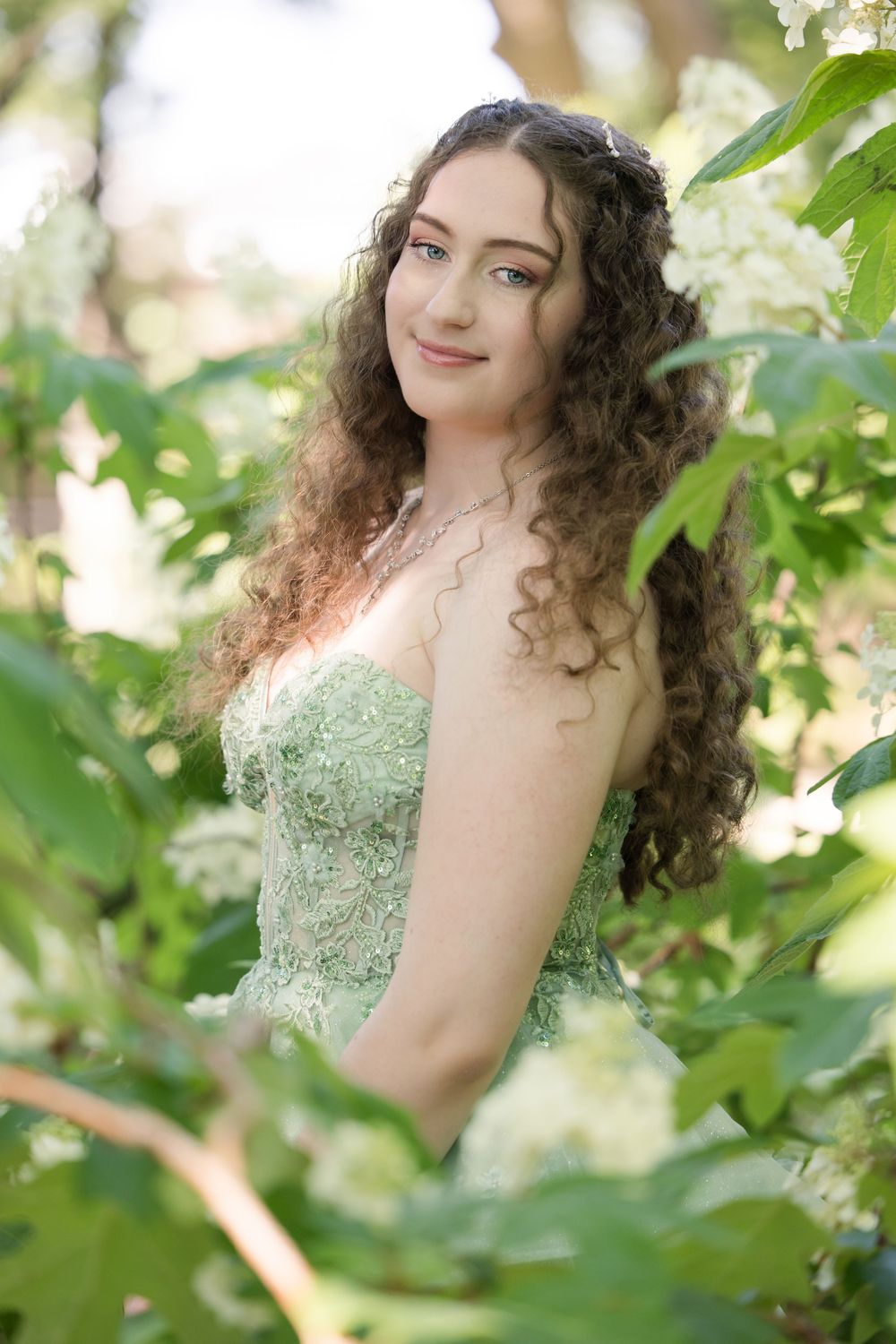 A portrait in nature showing curly long hair and a mint green lace dress among flowering white blossoms and green leaves.