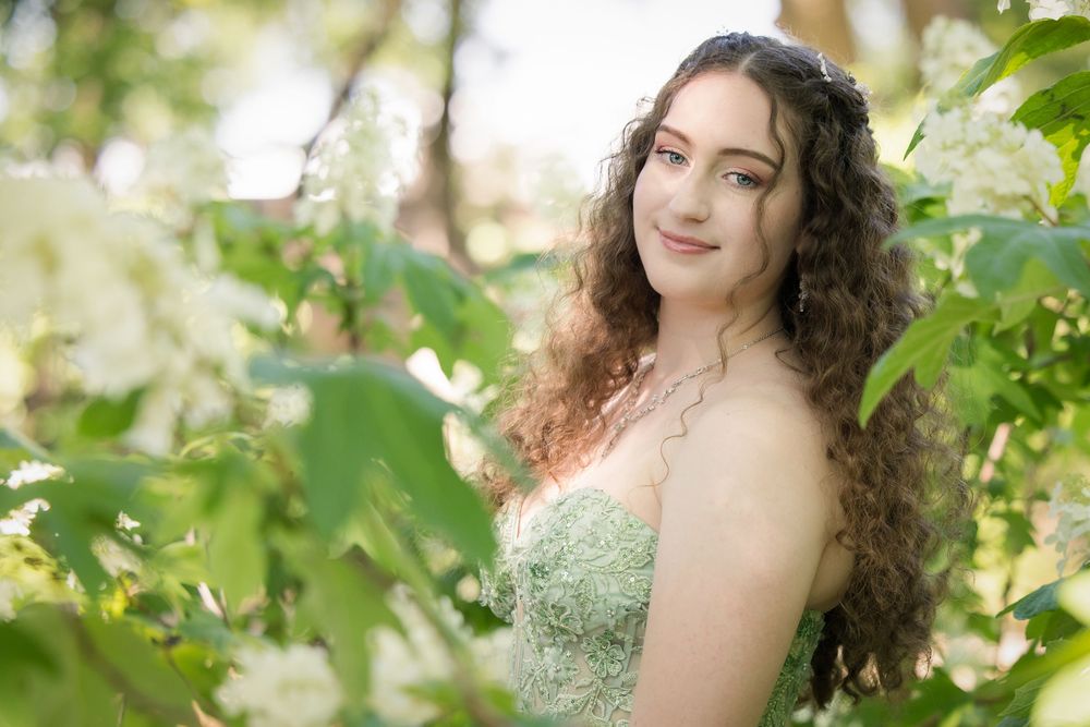 A curly-haired figure in a sparkly green dress poses among leafy branches in soft natural lighting.
