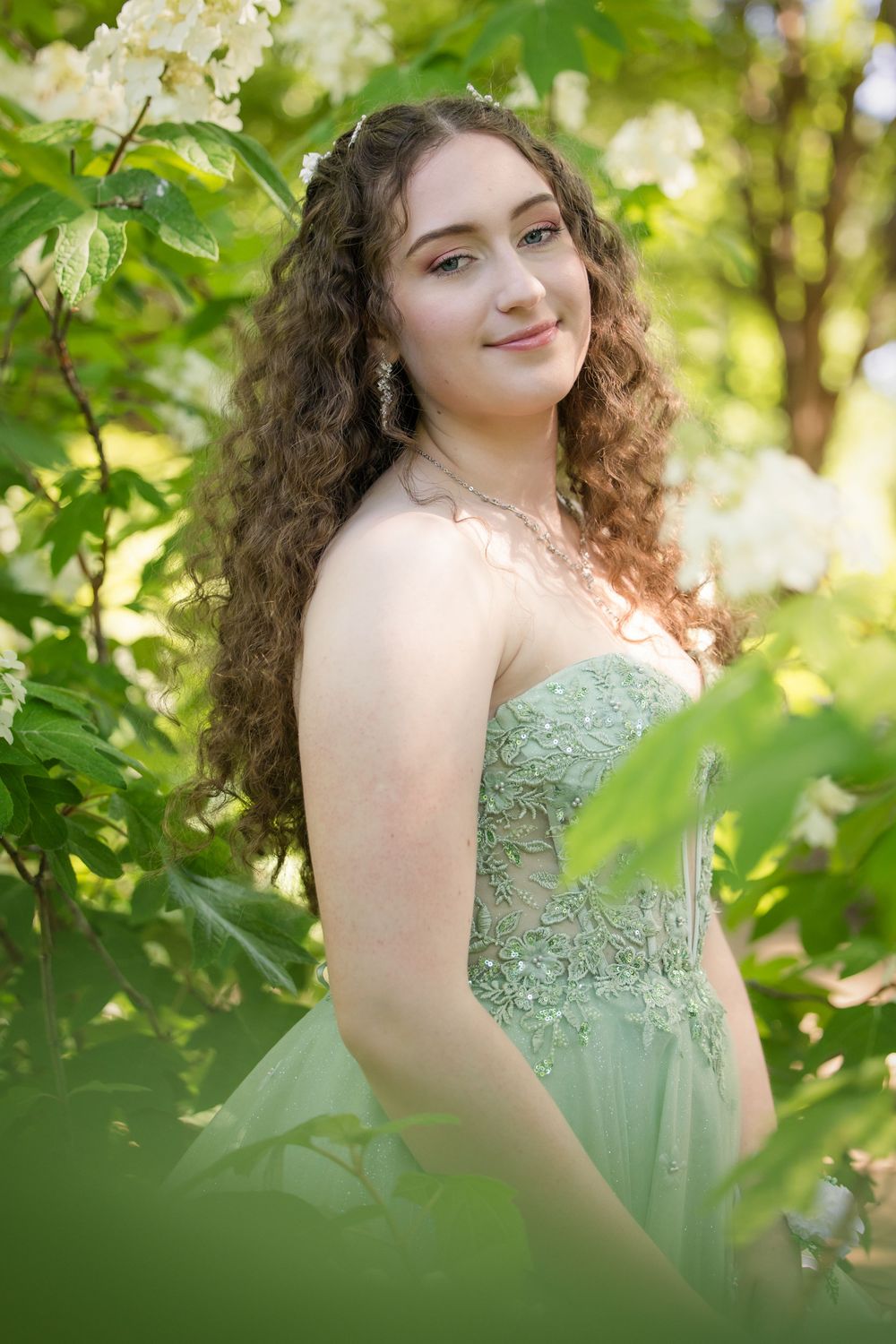 A young woman in a mint green formal dress poses among blooming spring foliage in a garden setting.