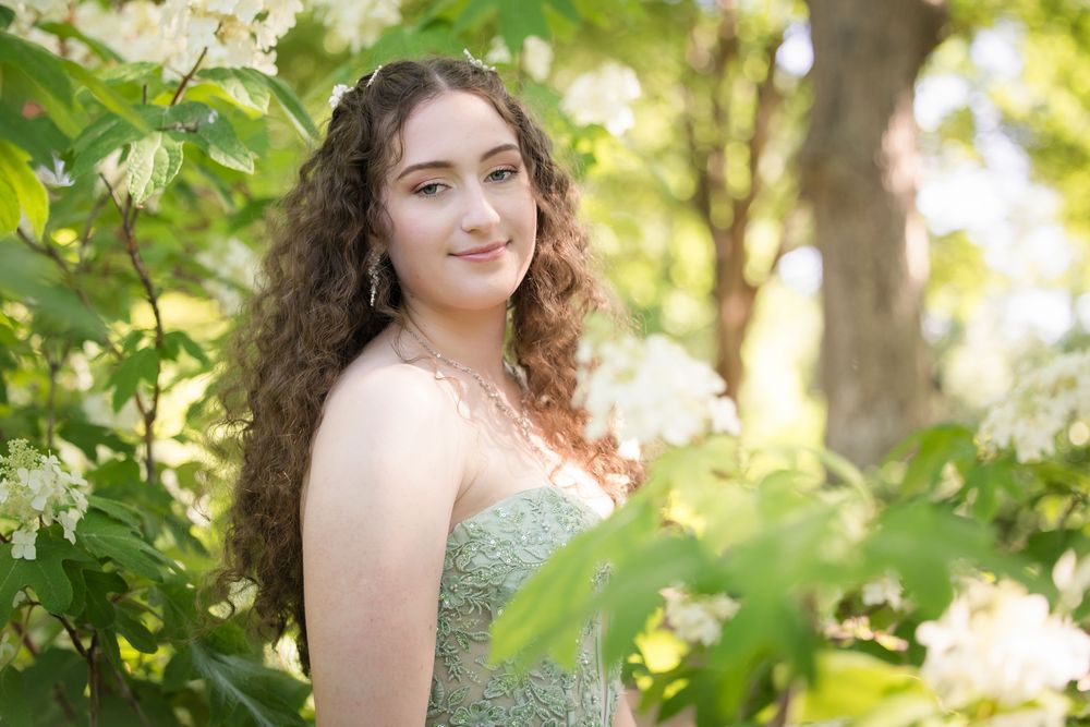A person in a mint green formal dress poses outdoors among green foliage in natural sunlight.