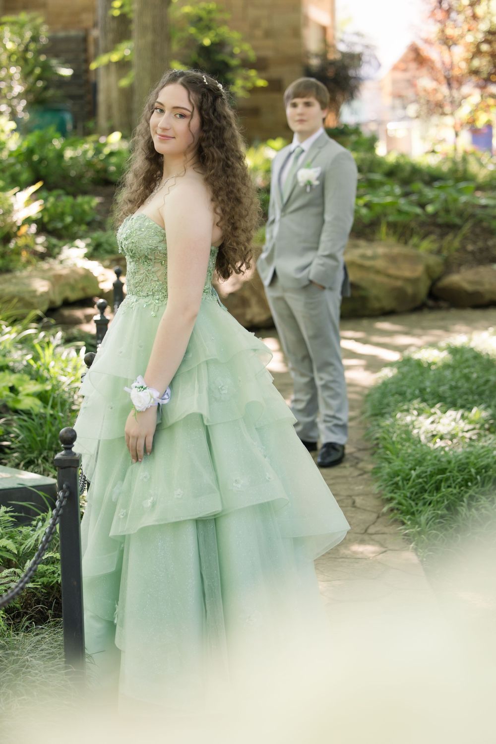 Formal couple poses in garden setting with woman in mint green layered prom dress and man in grey suit.