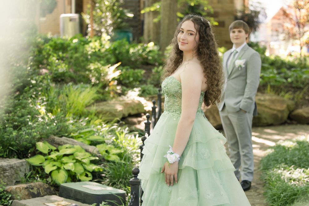 A person in a pale mint green prom dress poses in a garden while someone in a gray suit stands in the background.