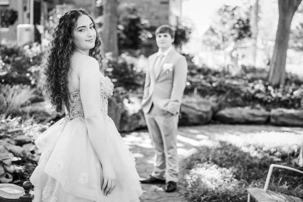 Black and white portrait of a bride in a flowing gown posing in a garden with another figure in the background.