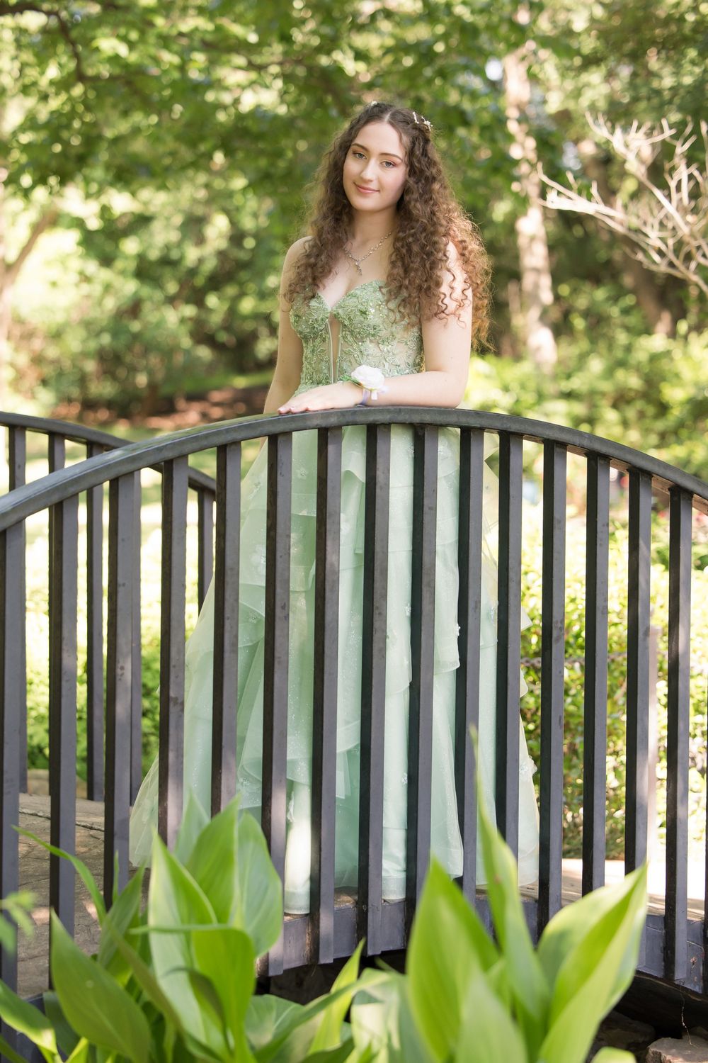 A person in a formal sage green dress poses on an arched garden bridge surrounded by lush summer foliage.