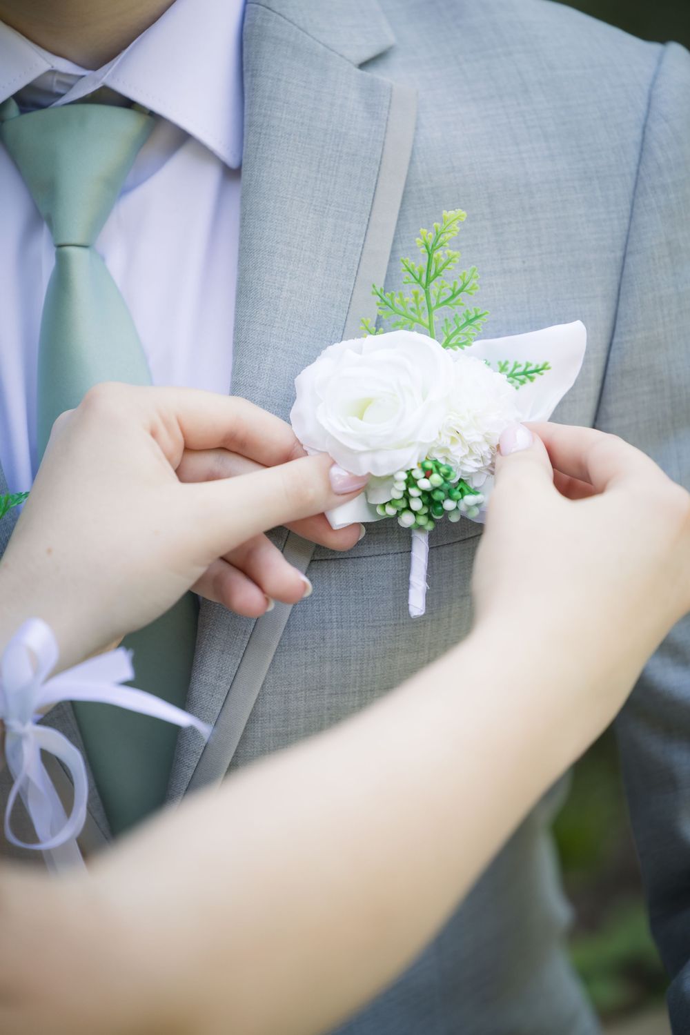 Hands pin a white rose and greenery boutonniere onto a light gray suit jacket with mint green tie.