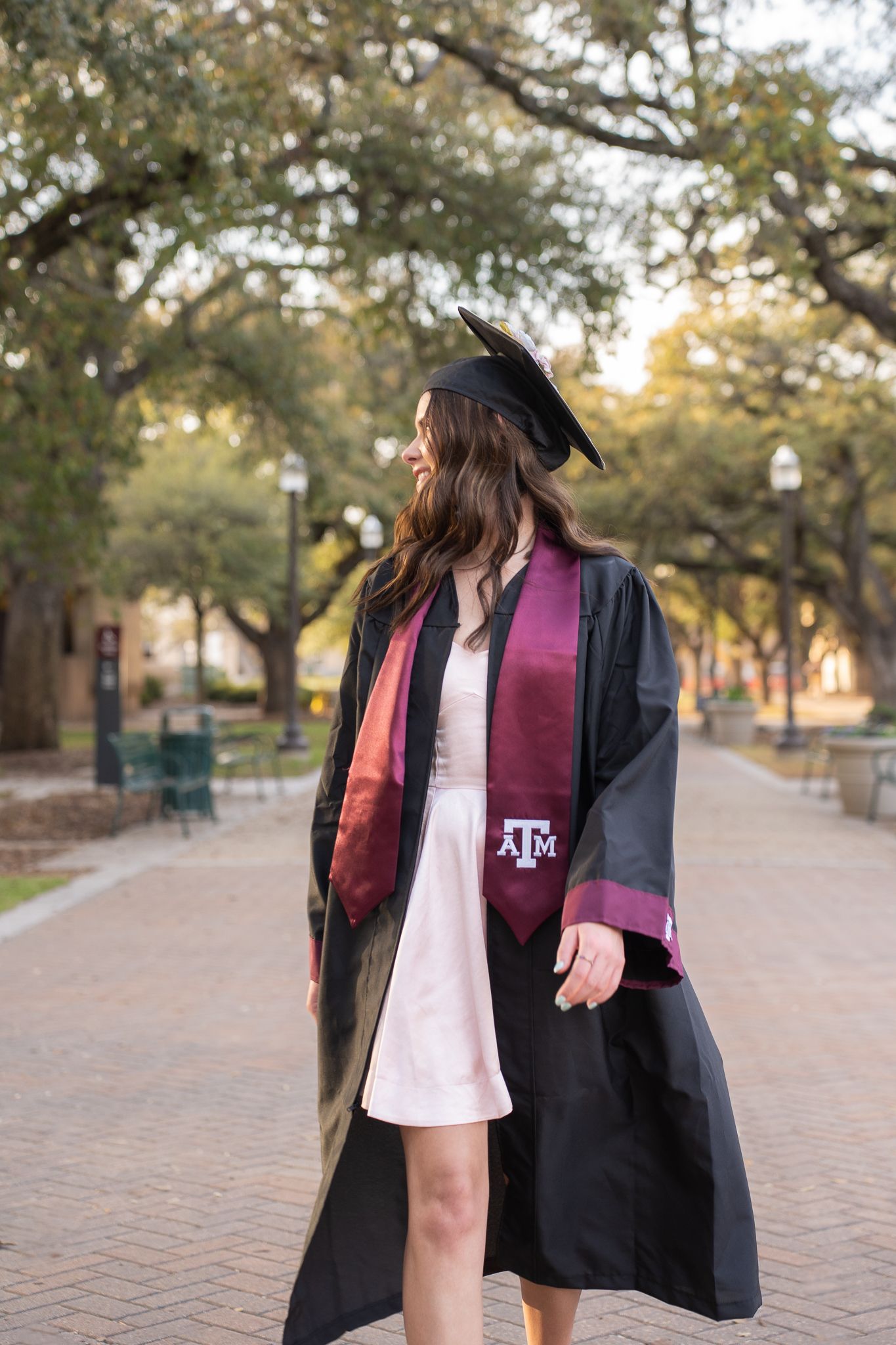 Graduate in black cap and gown with maroon Texas A&M stole walking on brick pathway surrounded by trees.