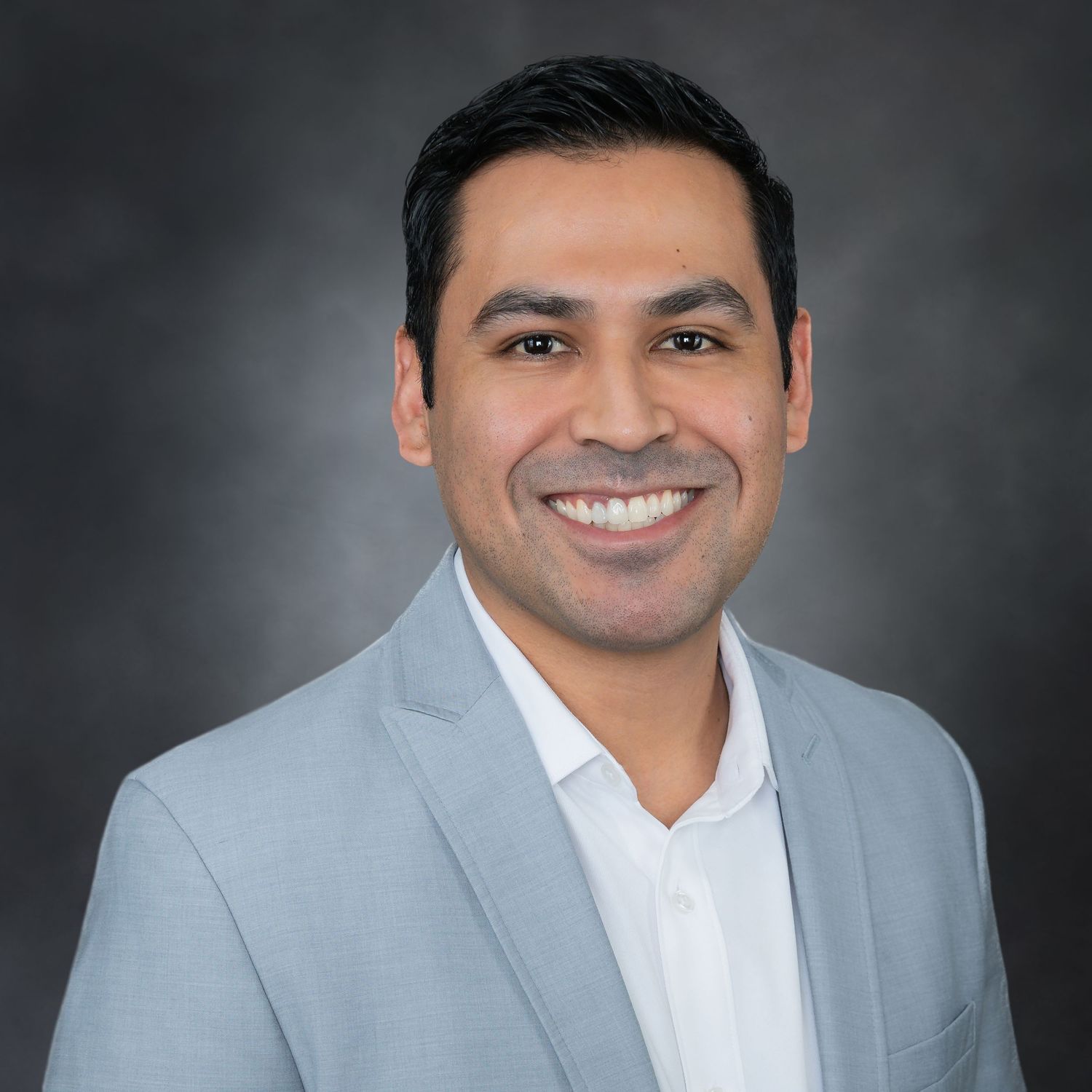 Professional headshot of person in grey suit jacket and white dress shirt against dark background with warm lighting.