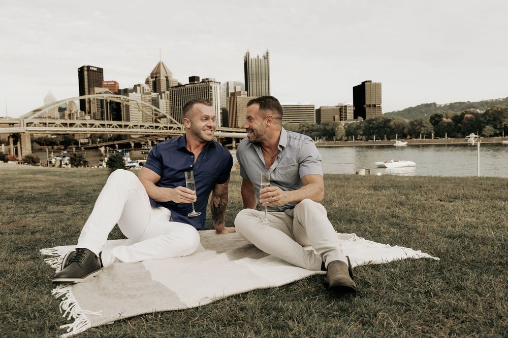 Two people enjoy drinks while relaxing on a blanket at Point State Park with the Pittsburgh skyline in background.