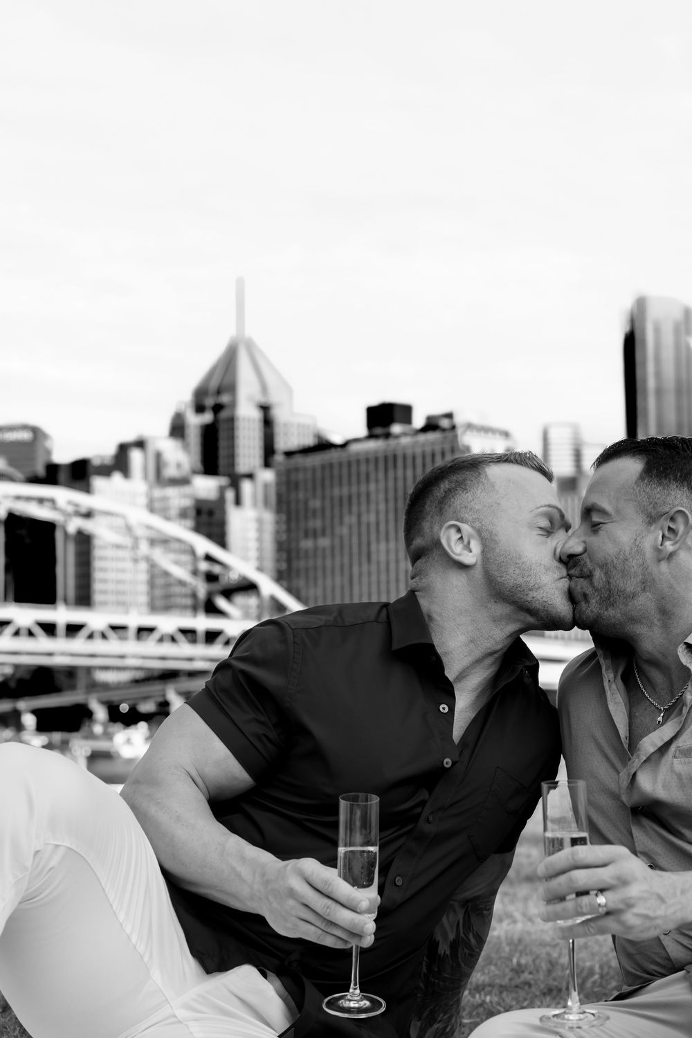 Two people sharing a romantic kiss outdoors with the Pittsburgh cityscape and iconic downtown buildings in the background.