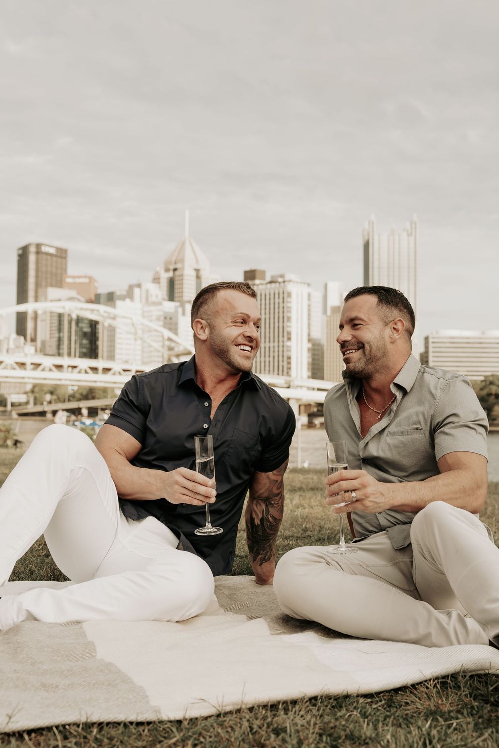 Two people enjoying drinks while sitting on a blanket with the Pittsburgh city skyline visible in the background.