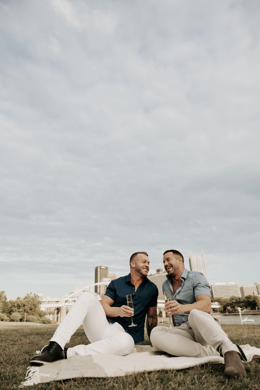 Two people enjoy drinks while sitting on a blanket in a park with an urban skyline in the background on a cloudy day.