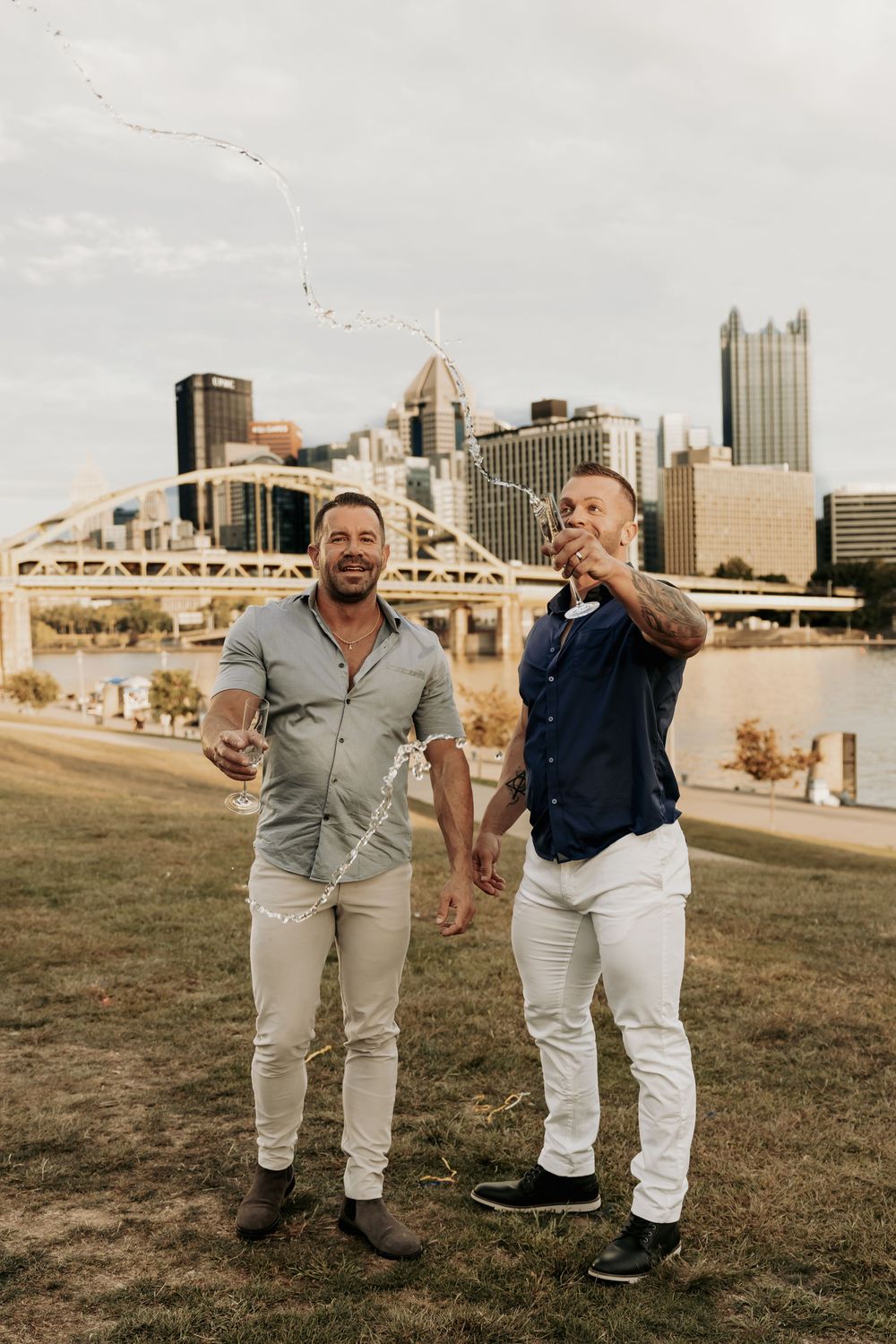 A couple wearing coordinating outfits stands on a grassy hill with the Pittsburgh skyline and bridges visible behind them.