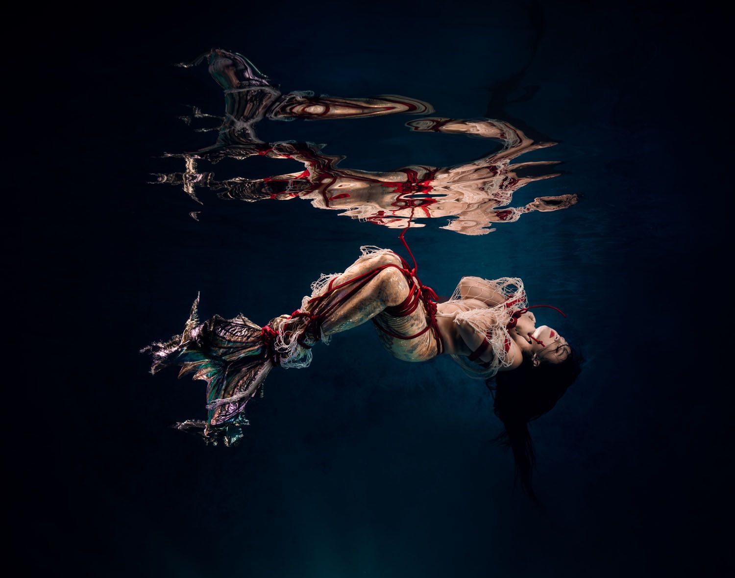 Underwater fashion photography captures a floating figure in a red outfit with flowing fabric against dark water.