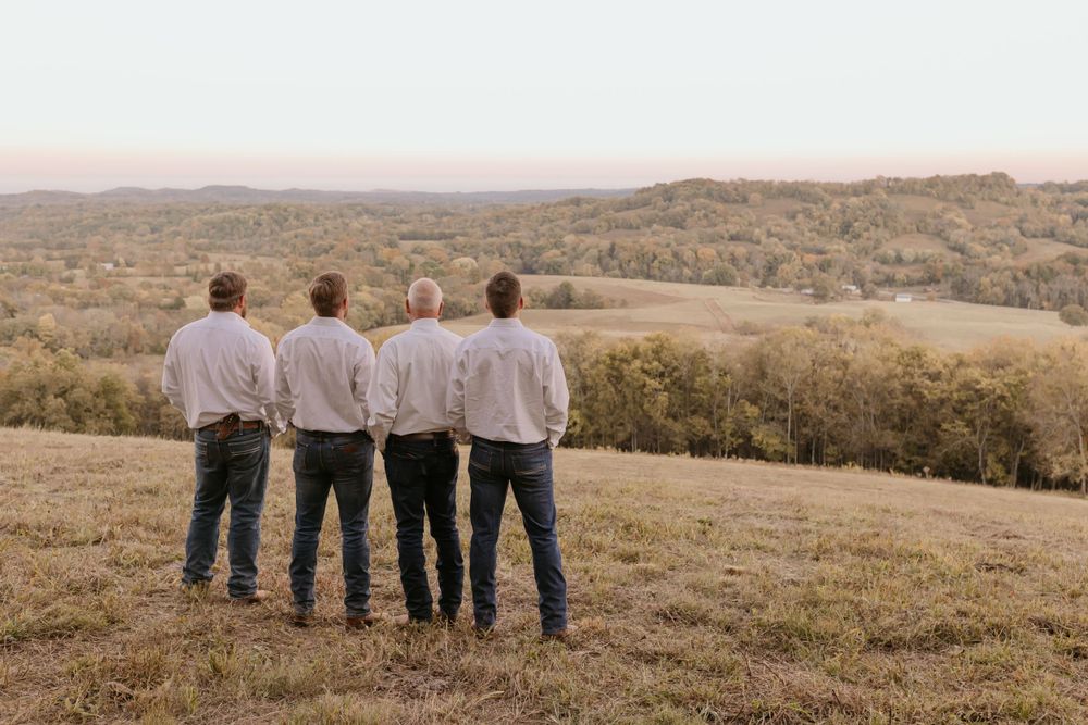 Four people stand on a hilltop overlooking a scenic autumn landscape at sunset.