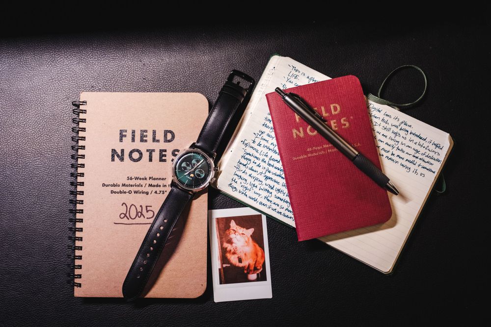 Travel essentials including Field Notes notebook, passport, polaroid photo and wristwatch on dark surface.