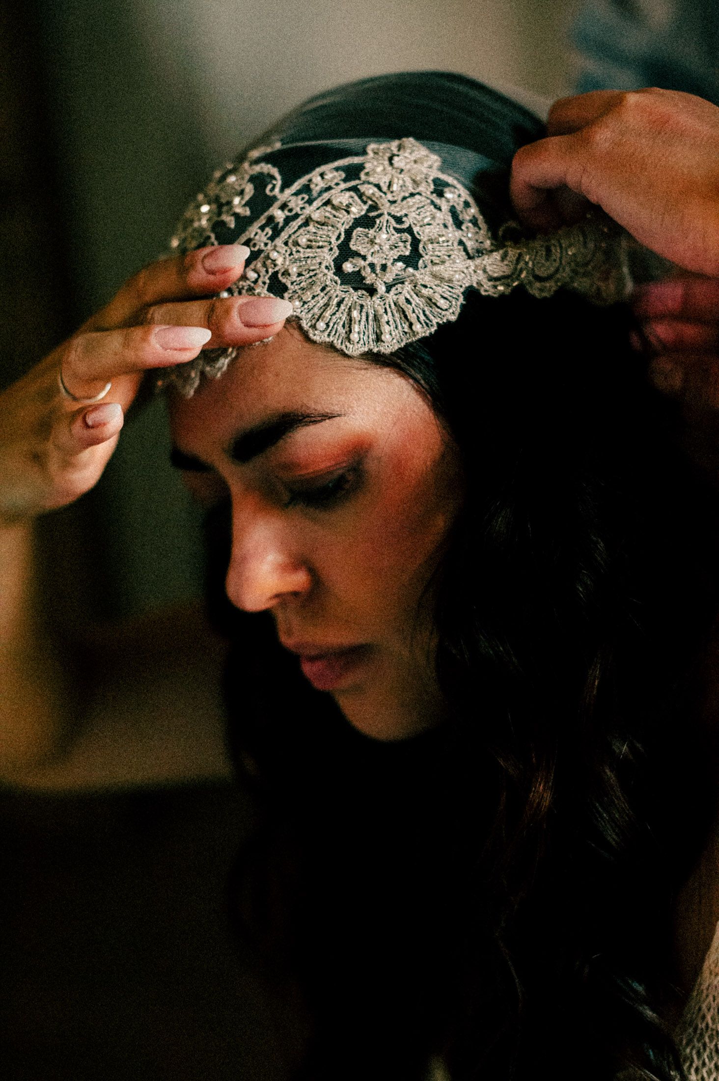 Intricate beaded headpiece being placed on dark-haired woman in romantic lighting.