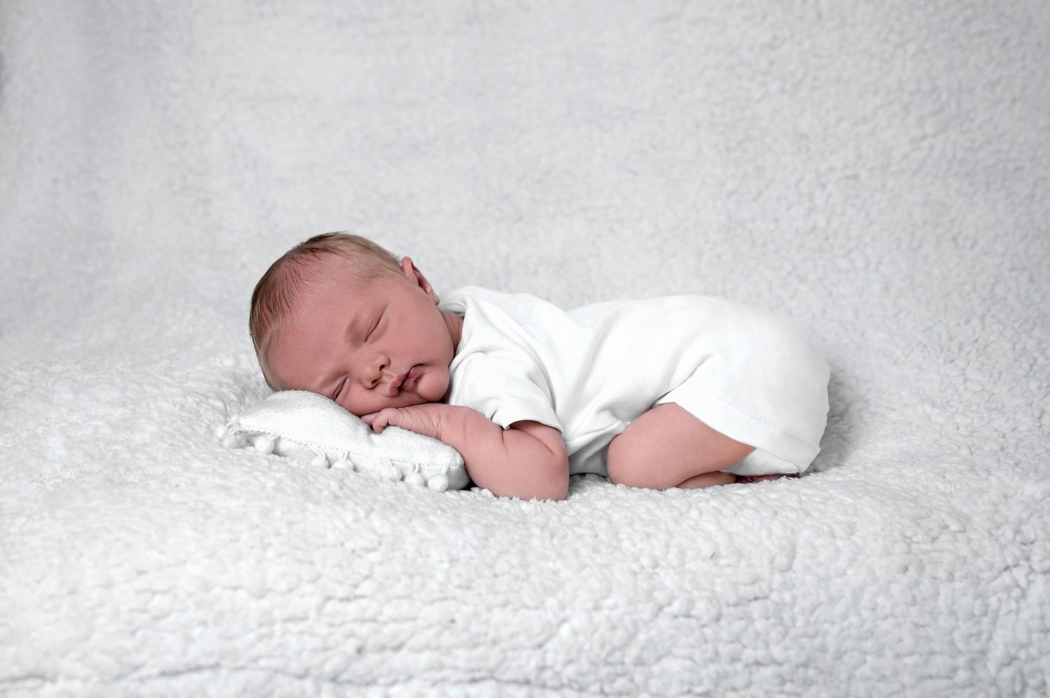 A sleeping newborn baby dressed in white lies peacefully on a soft white textured blanket.