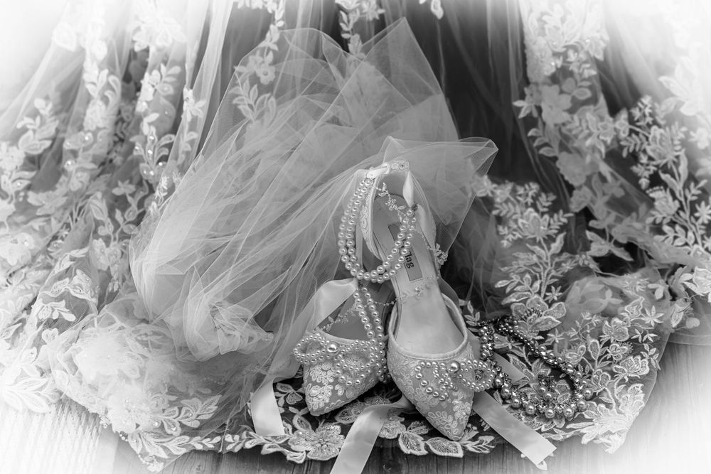 Elegant bridal accessories featuring pearl-adorned heels and delicate lace veil arranged on white floral lace background.