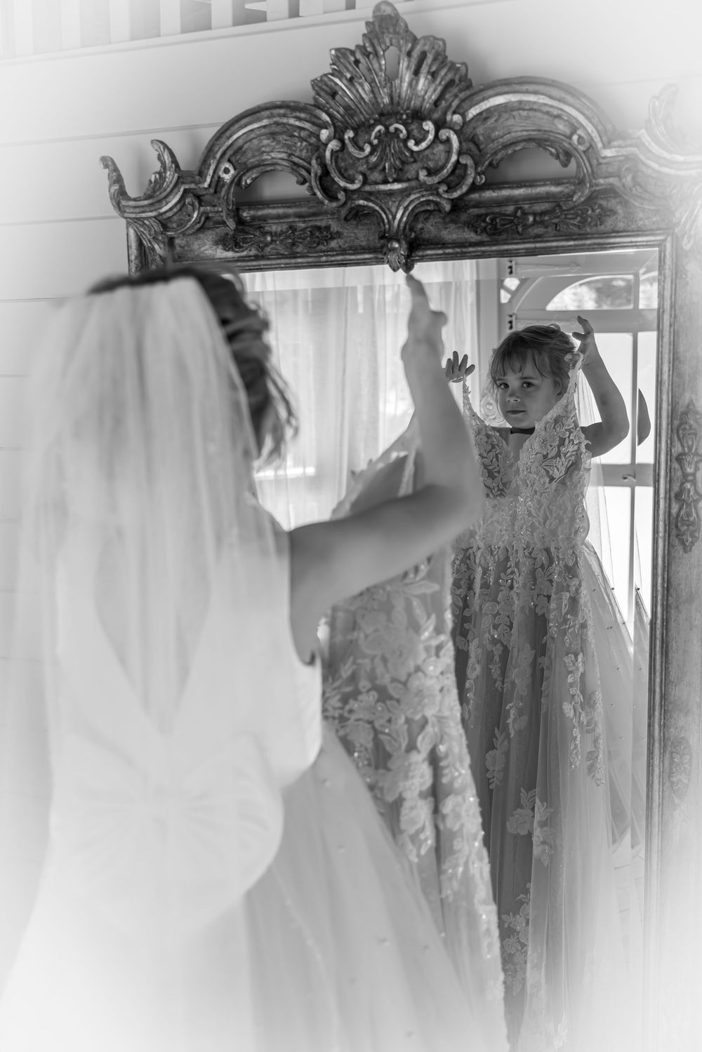 Black and white vintage photo showing lace wedding gown reflection in an ornate mirror with decorative carved frame.
