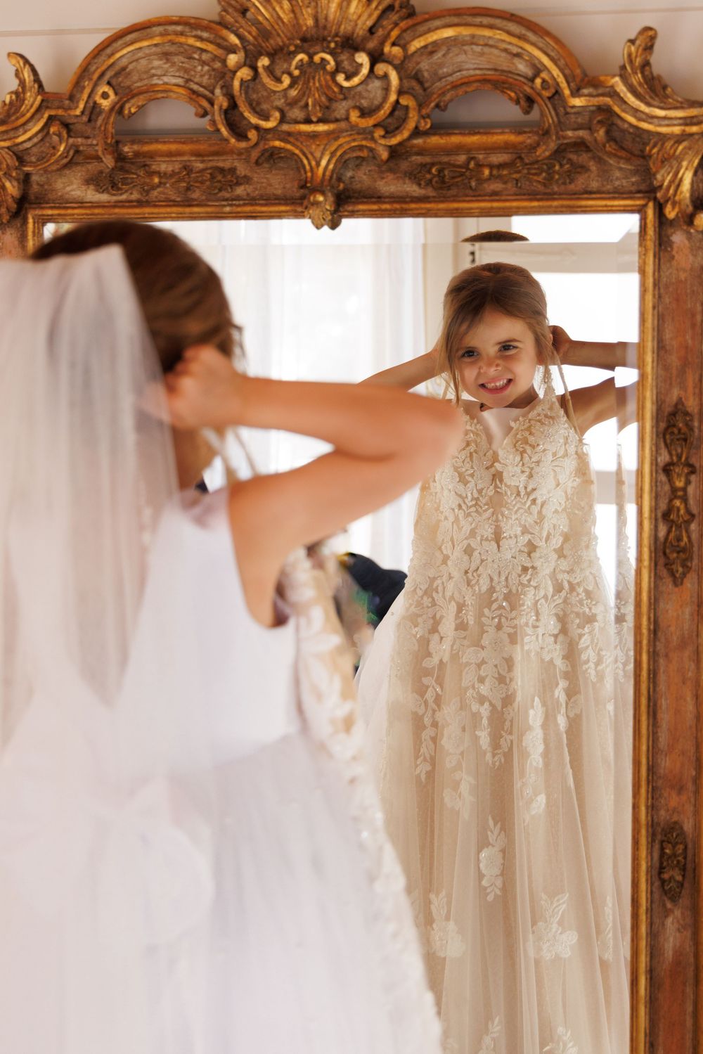 A person in a wedding dress admires their reflection in an ornate gold antique mirror while wearing a delicate lace gown.