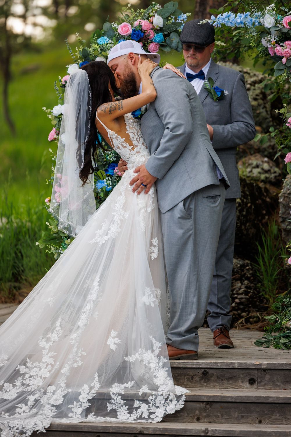 A wedding couple shares a romantic kiss on garden stairs framed by pink and blue flowers with an officiant in the background.
