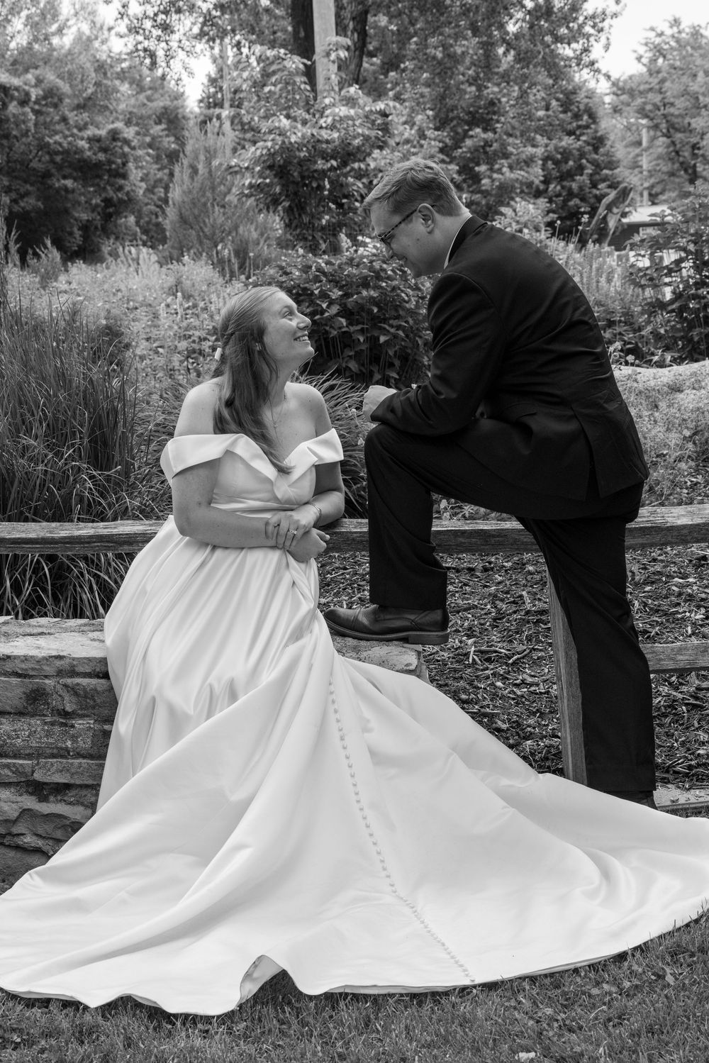 A black and white photo of two people in elegant wedding attire sitting together on a garden wall by greenery.