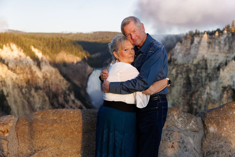 A couple embraces while standing against a majestic mountain cliff backdrop at sunset.