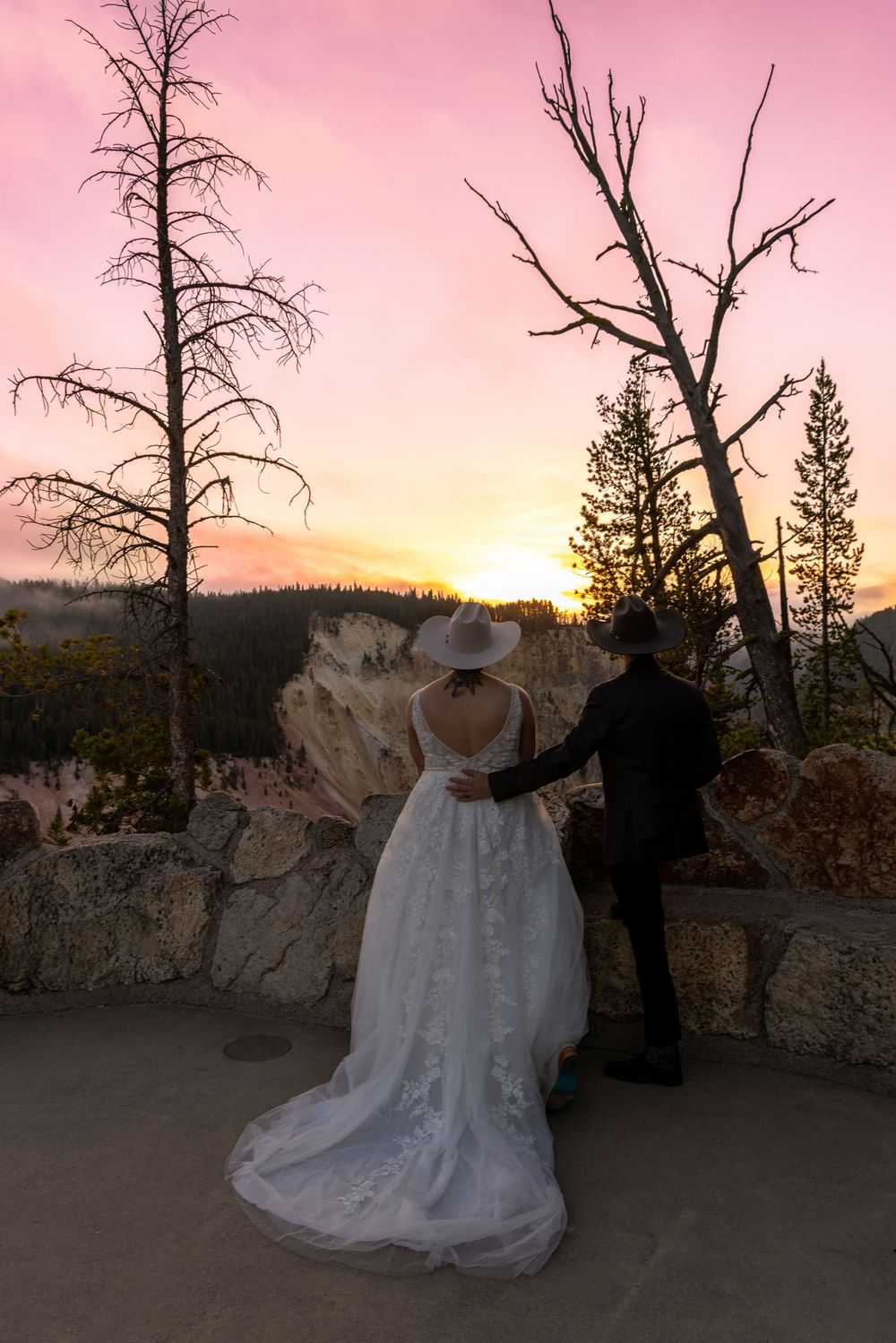 A wedding couple admires a vibrant pink and orange sunset from a scenic mountain viewpoint.